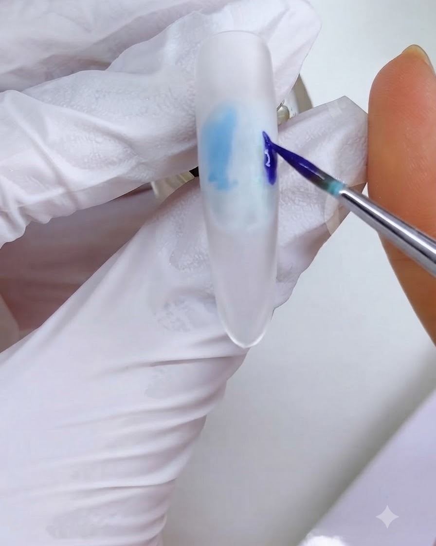 Ein feiner Liner-Pinsel trägt einen blau-lila Tintenblock auf die rechte Seite der weiß gefrosteten Nageloberfläche auf.