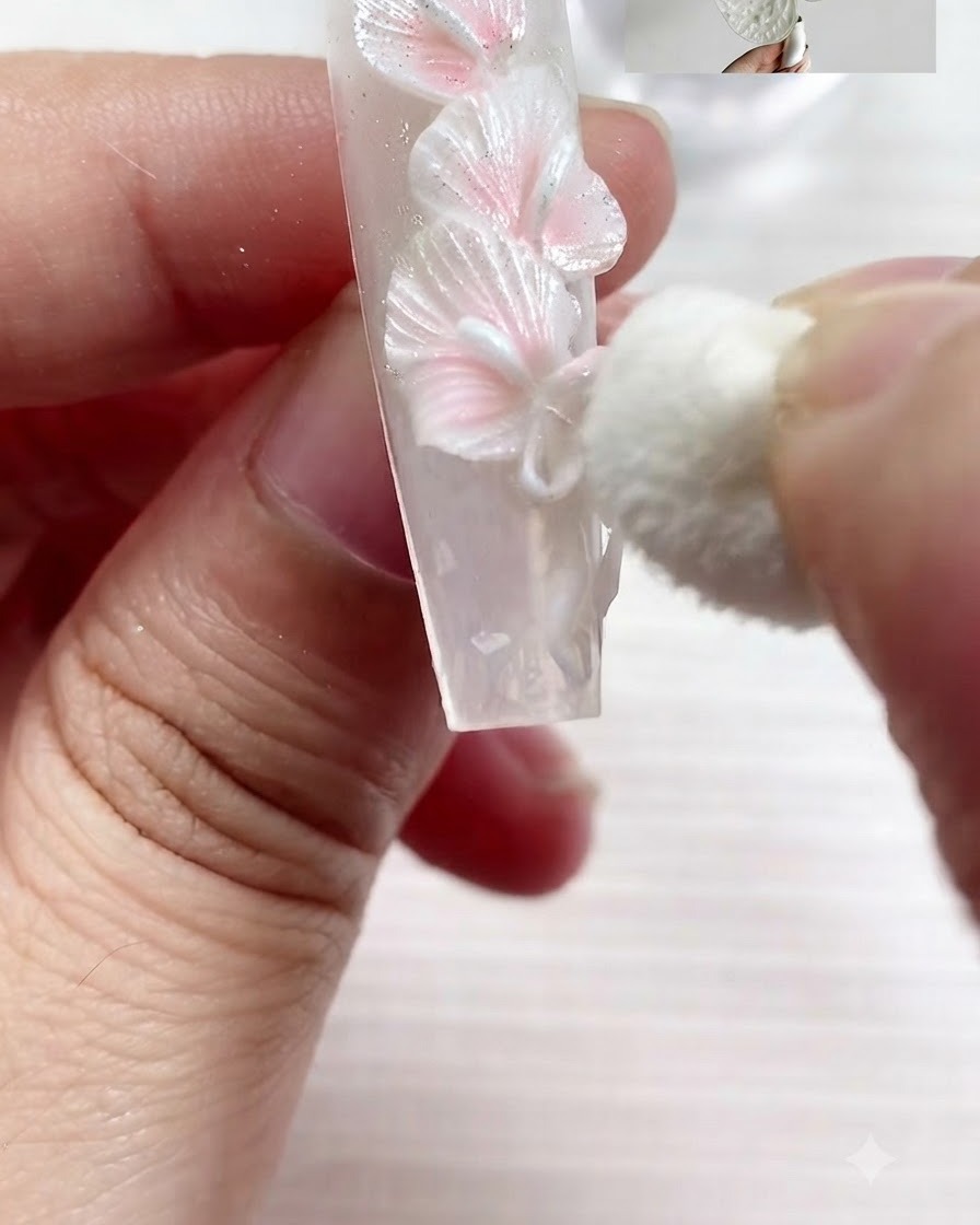 Ein kleiner Make-up-Schwamm tupft sanft Perlweiß-Pigment auf die strukturierten Kanten der skulpturierten weißen Calla-Lilien und einer Schleife auf einem Nagel.