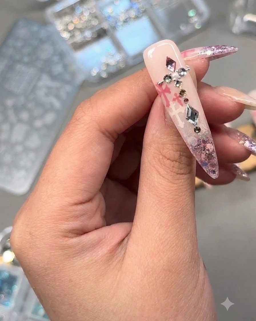 Nahaufnahme eines verzierten Nagels mit Kristallen, einer Perle und Metall-Charms über rosa Glitzer und Stempeling-Art.