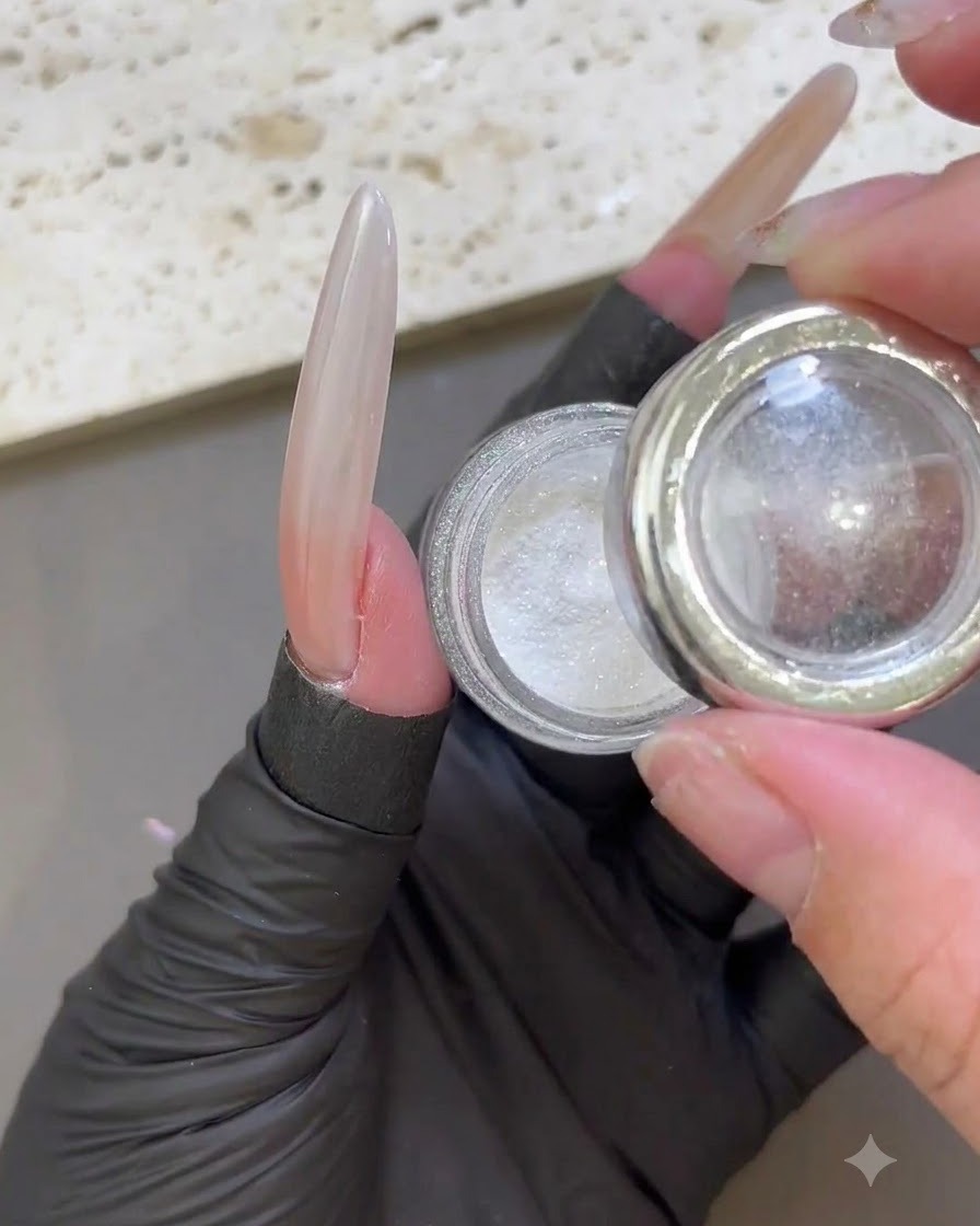 Eine handschuhtragende Hand hält ein geöffnetes Glas mit weißem, irisierendem Moonlight-Puder neben einen glänzenden verlängerten Nagel.