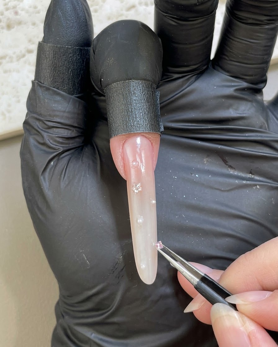 Eine handschuhtragende Hand verwendet eine Pinzette, um einen kleinen rosa Strassstein auf eine verzierte Nageloberfläche zu setzen.