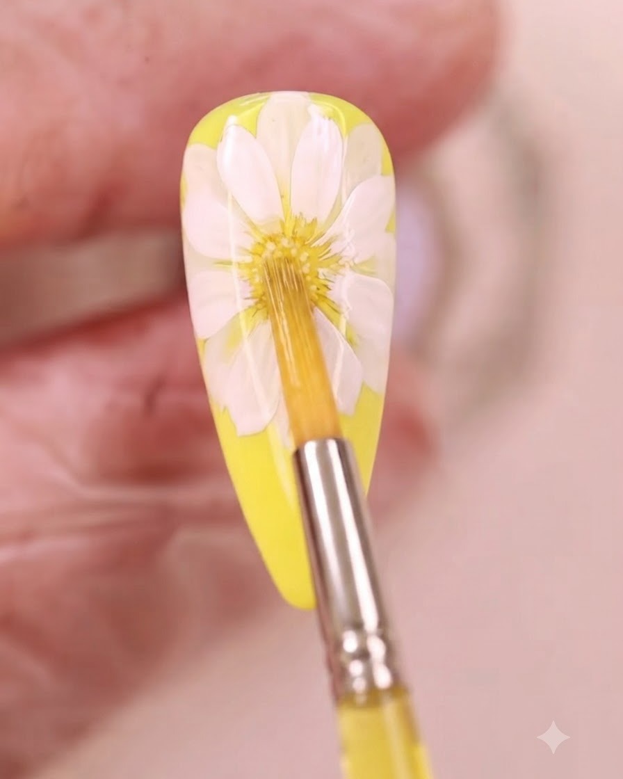 Ein feiner Pinsel trägt gelbe Aquarellflüssigkeit auf die Mitte eines weißen Gänseblümchen-Nageldesigns auf.