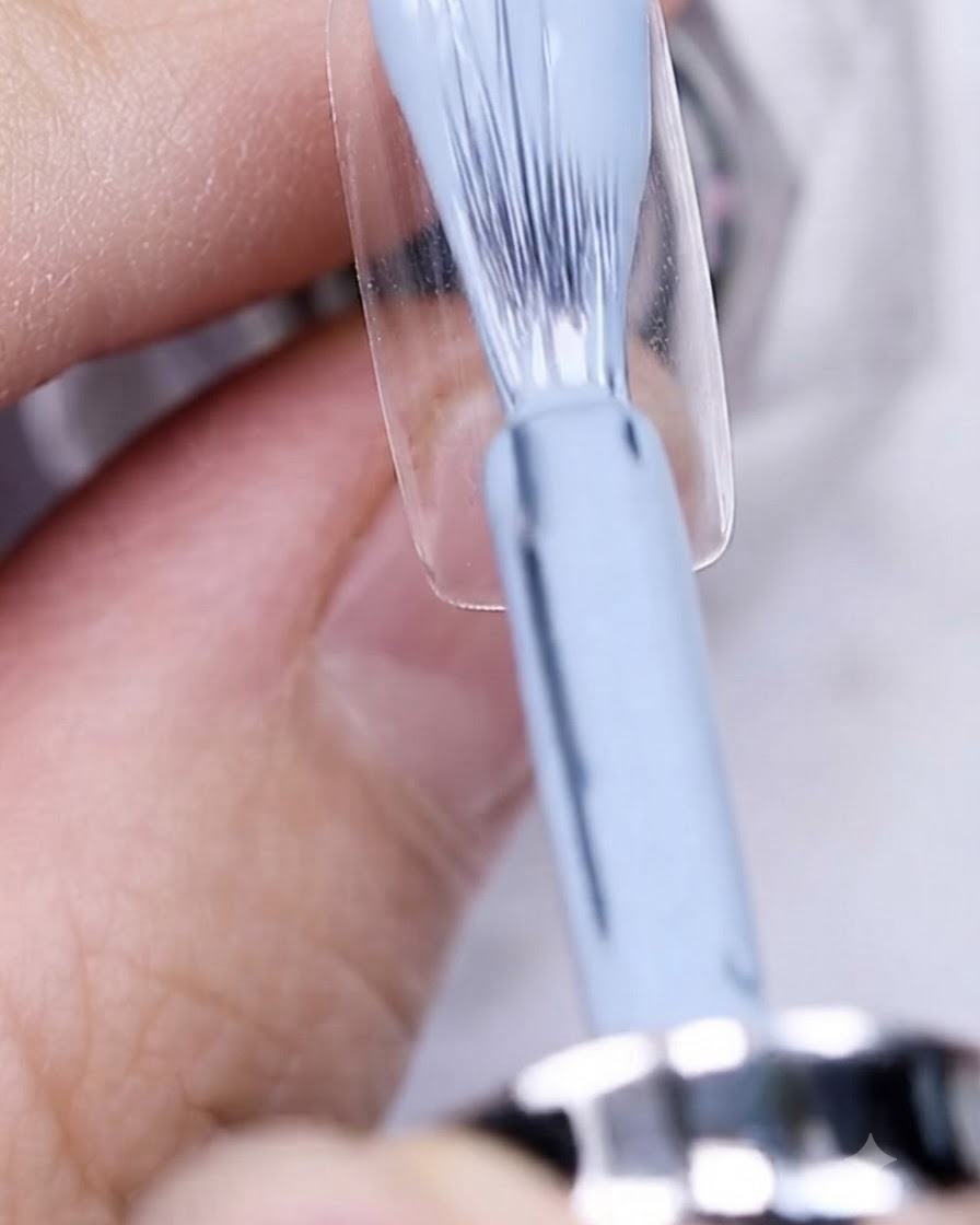 Ein Pinsel trägt graublauen Gellack auf eine unlackierte Nagelplatte auf.