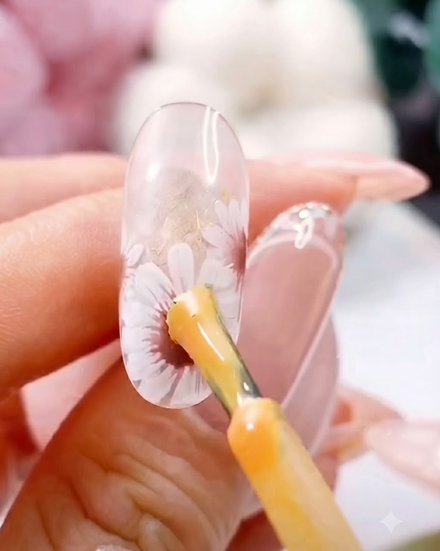 Ein Pinsel trägt einen dicken gelben Gelpunkt auf die Mitte eines gemalten weißen Gänseblümchens auf einem klaren Nagel auf.