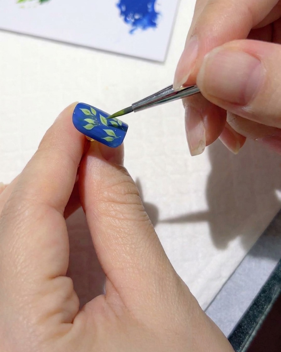 Ein feiner Nail-Art-Pinsel, der hellgrüne Striche über dunkle Blätter auf einem blauen Nagel aufträgt.