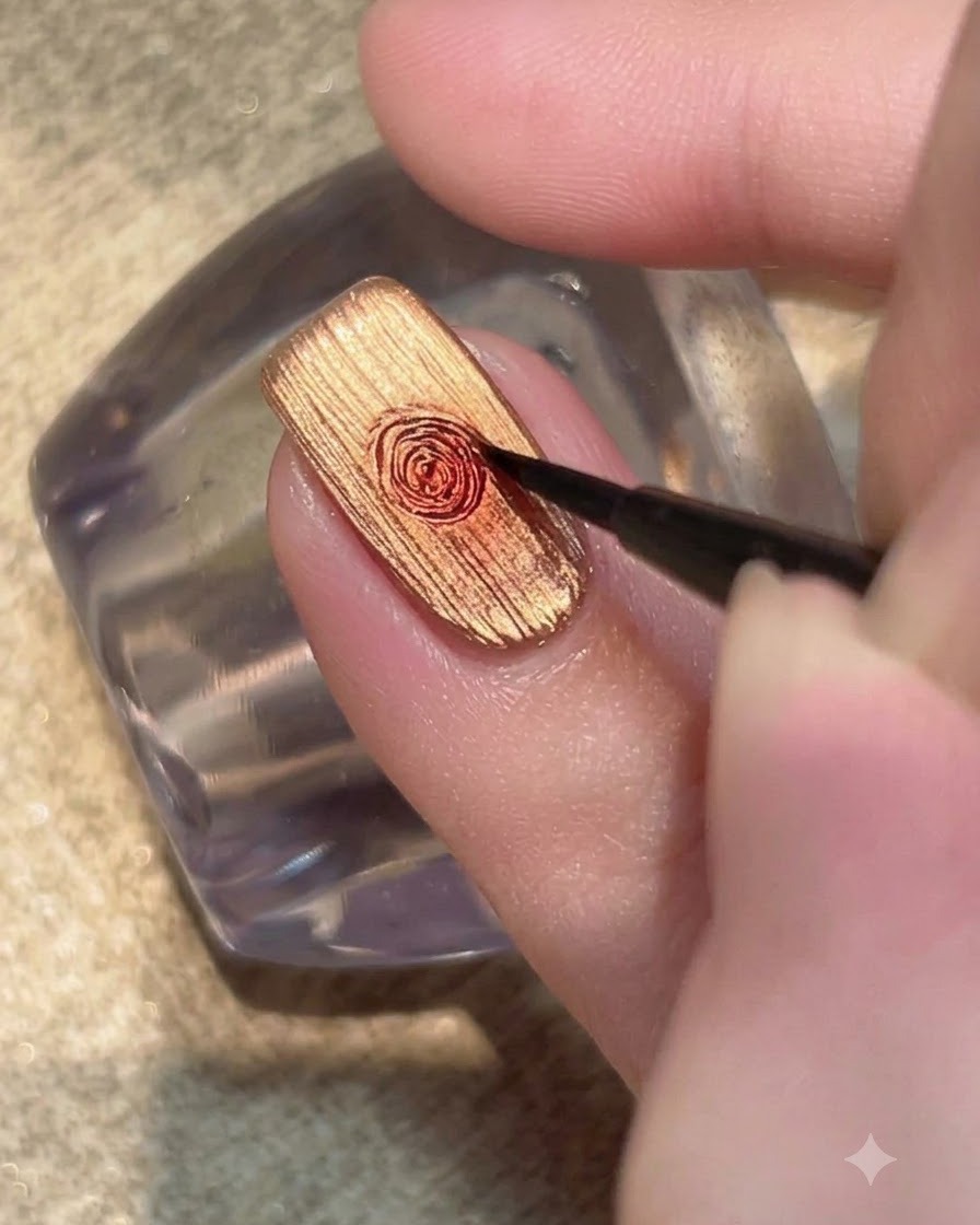 Ein feiner Detailpinsel verblendet einen Tropfen rotes Gel zu einem kreisförmigen Rosenmuster auf einem metallisch goldenen Nagel.