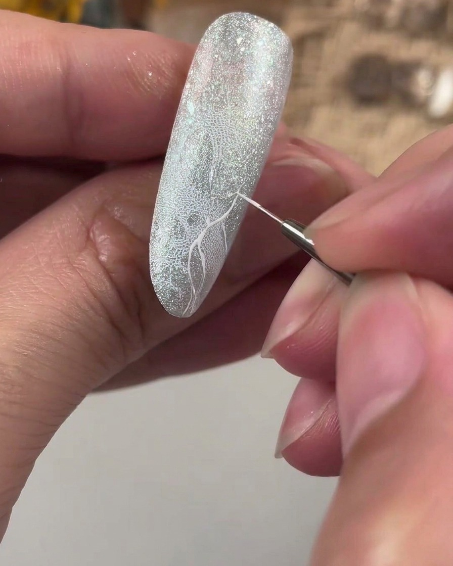 Ein feiner Detailpinsel zeichnet ein weißes Hilfslinienkreuz auf einen strukturierten Glitzernagel.