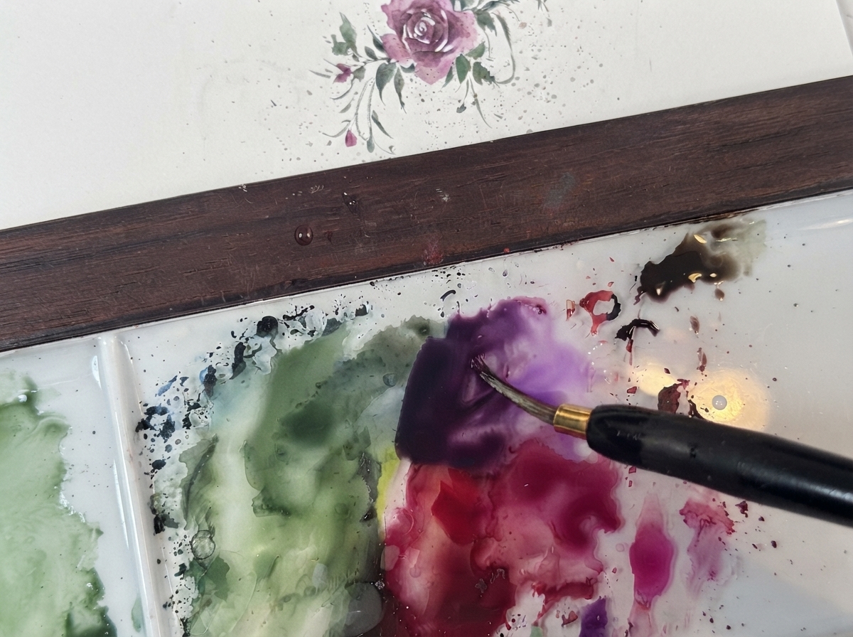Ein feiner Aquarellpinsel mischt dunkelviolettes Pigment auf einer sauberen weißen Palette neben grünen Lasuren.