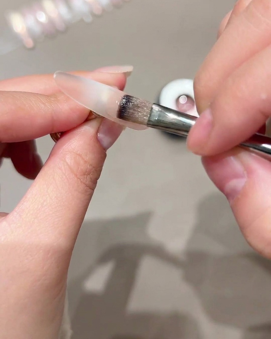 Ein Pinsel trägt Cat-Eye-Gel auf den äußeren Teil einer durchsichtigen Nagelspitze auf.