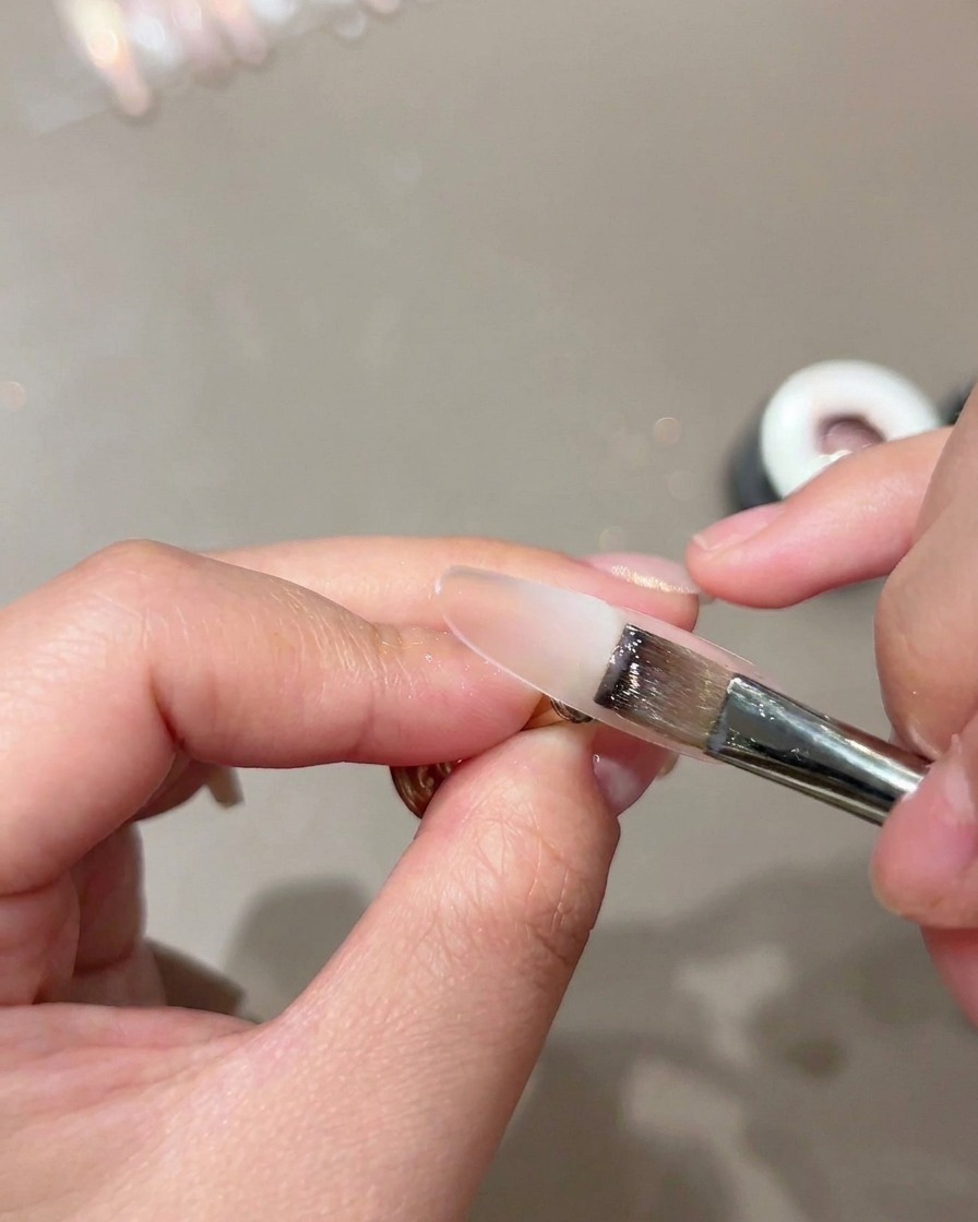 Ein flacher Pinsel trägt eine transparente Nude-Basisschicht auf eine Nagelform auf.