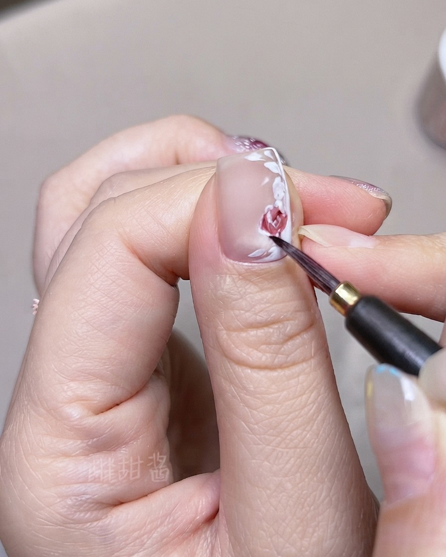 Ein feiner Pinsel schichtet dunklere rote Aquarelltöne auf individuell gemalte Rosenblüten und Blätter auf einem Fingernagel.