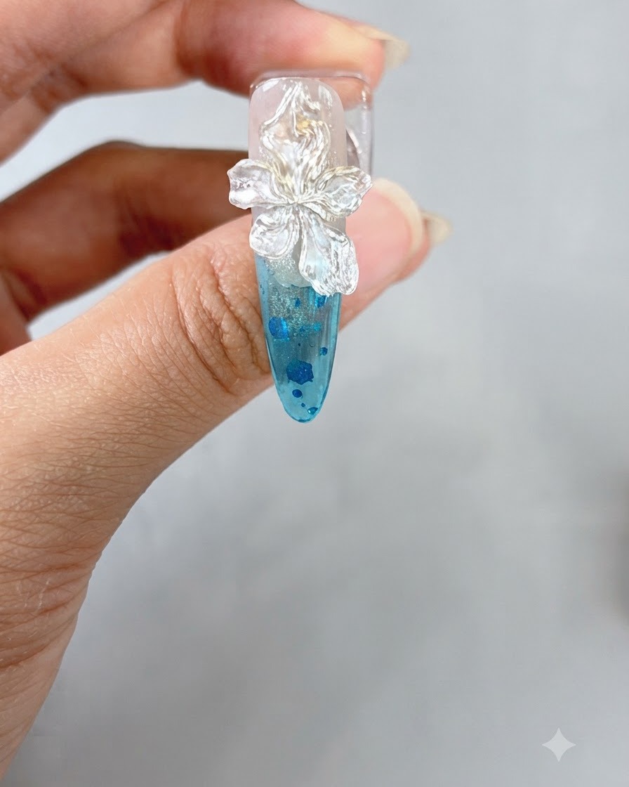Eine Pinzette platziert eine kleine weiße Perle auf die Mitte einer skulpturierten 3D-Blume auf einem Nagel.
