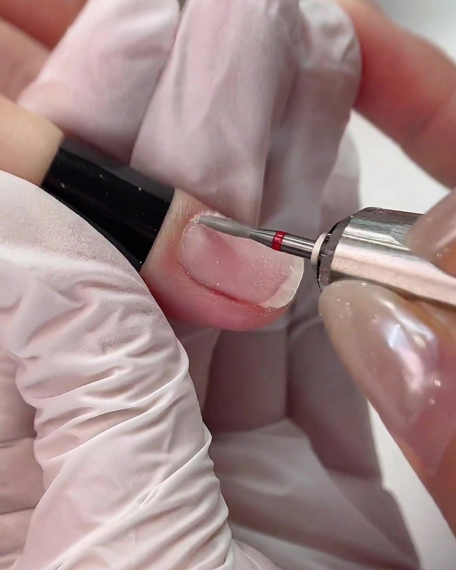 Eine Fräse mit einem rot markierten Diamant-Fräser arbeitet vorsichtig entlang der Nagelhautlinie eines Fingers.