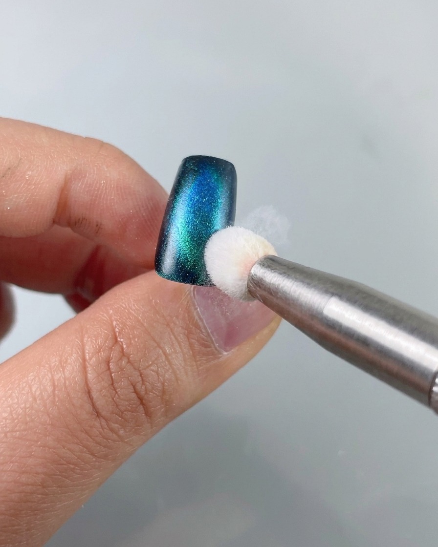 Ein Schwammwerkzeug tupft schwarzes Stamping-Gel auf den Rand eines blau-grünen Cat-Eye-Nagelstücks.