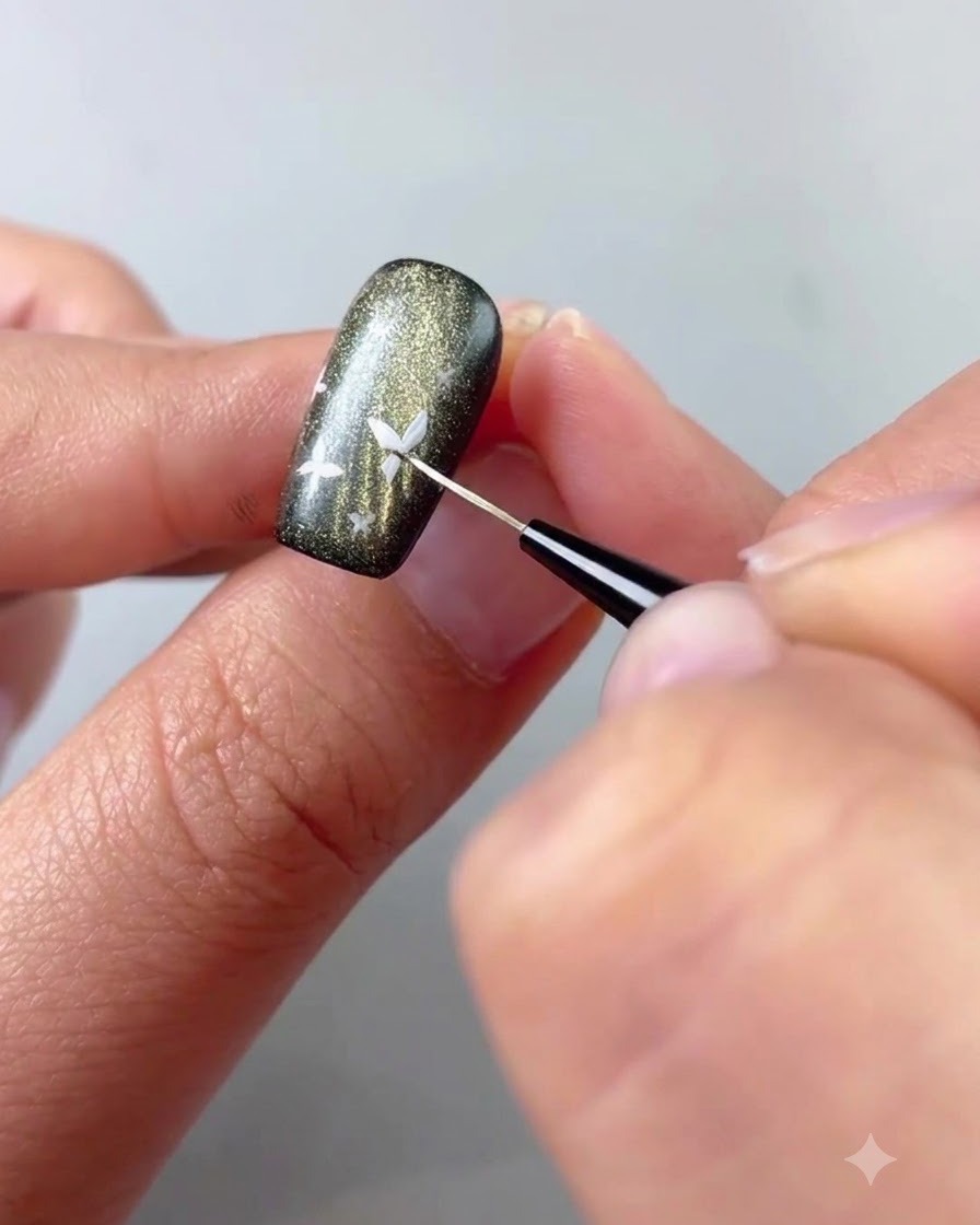 Eine Nahaufnahme eines feinen Liner-Pinsels, der weißes Gel aufträgt, um ein kleines Schmetterlingsdetail auf einem dunklen Cat-Eye-Nagel zu malen.
