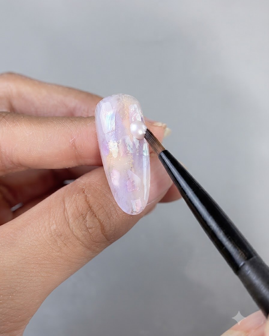 Ein feiner Kunstpinsel trägt einen Tropfen schimmerndes Gel auf eine verzierte, mit Muscheln besetzte Nageloberfläche auf.