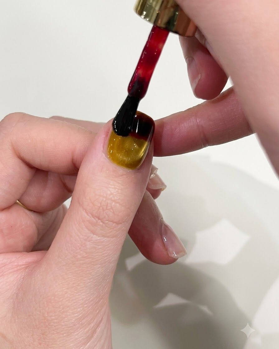Ein Nagellackpinsel trägt rotes Gel auf die obere Kante eines gelb manikürten Cat-Eye-Nagels auf.