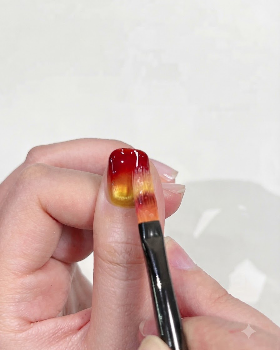 Ein flacher Nagelpinsel verblendet vorsichtig die rote Spitze nach unten in die gelbe Basis, um den Farbverlauf zu verfeinern.