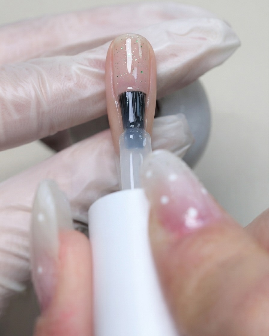 Ein Pinsel trägt klares C7 Flexibel-Base-Gel über die Nageloberfläche auf, um die platzierten Pailletten zu fixieren.