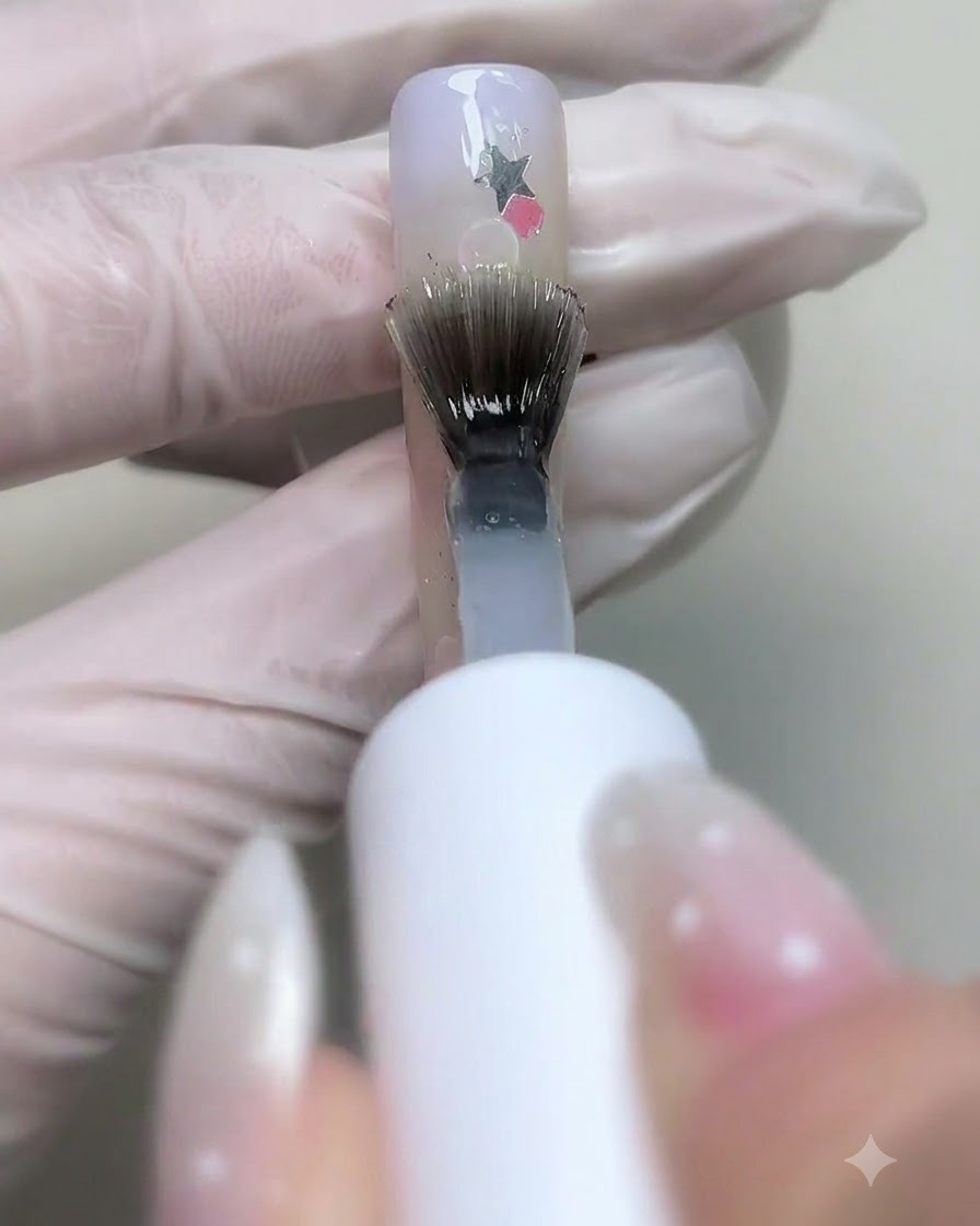 Ein Pinsel trägt klares Base-Gel über verzierte Nägel mit Sternen-Pailletten und Blasen auf.