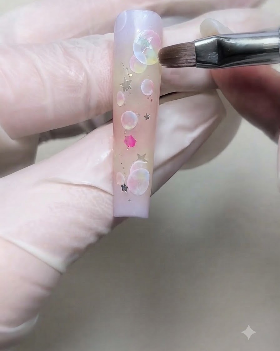 Eine bemalte Hand mit Handschuh platziert eine silberne Sternen-Paillette mit einem feinen Pinsel auf einem Gradienten-Nagel.
