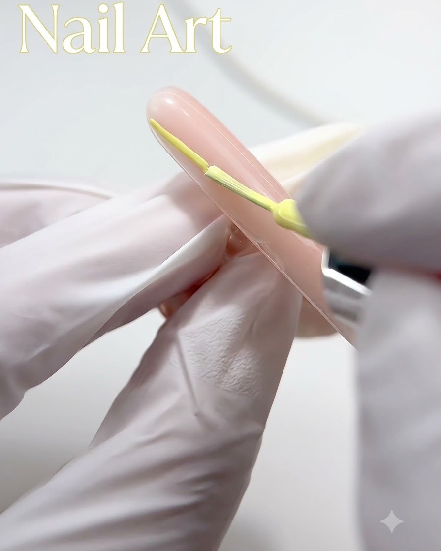 Eine handgeschützte Hand mit einem feinen Pinsel, die gelbes Gel-Polish auf die Spitze eines nudefarbenen Nagels aufträgt.