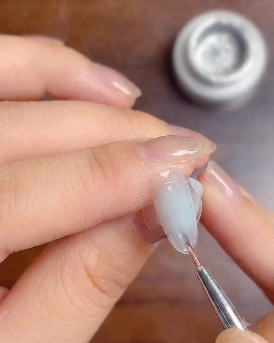 Ein feiner Pinsel trägt eine Schicht klaren Top-Coat-Gels über die Silberglitzerlinien auf einem Milky-Gradient-Nagel auf.