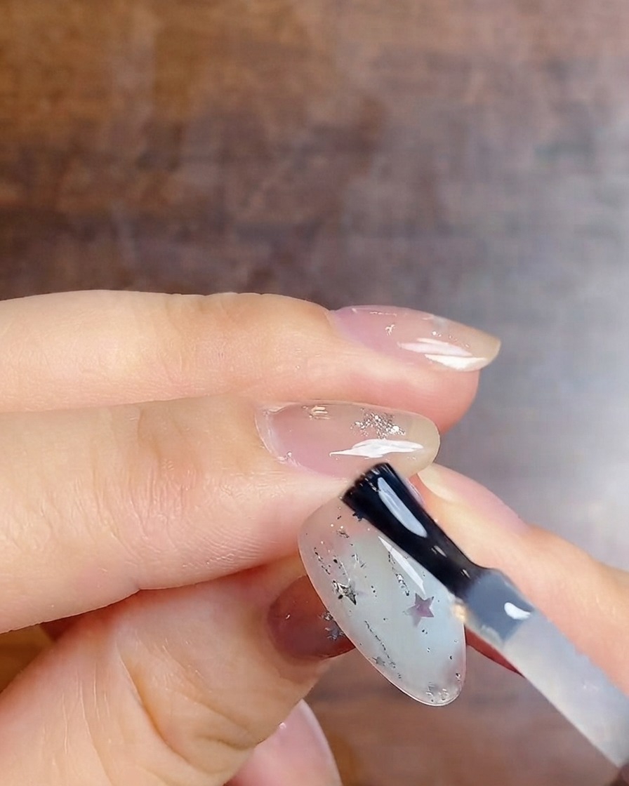 Ein Nagelpinsel trägt einen finalen glänzenden Top-Coat über einen Milchverlaufs-Nagel mit Silberglitzer und Stern-Stickern auf.