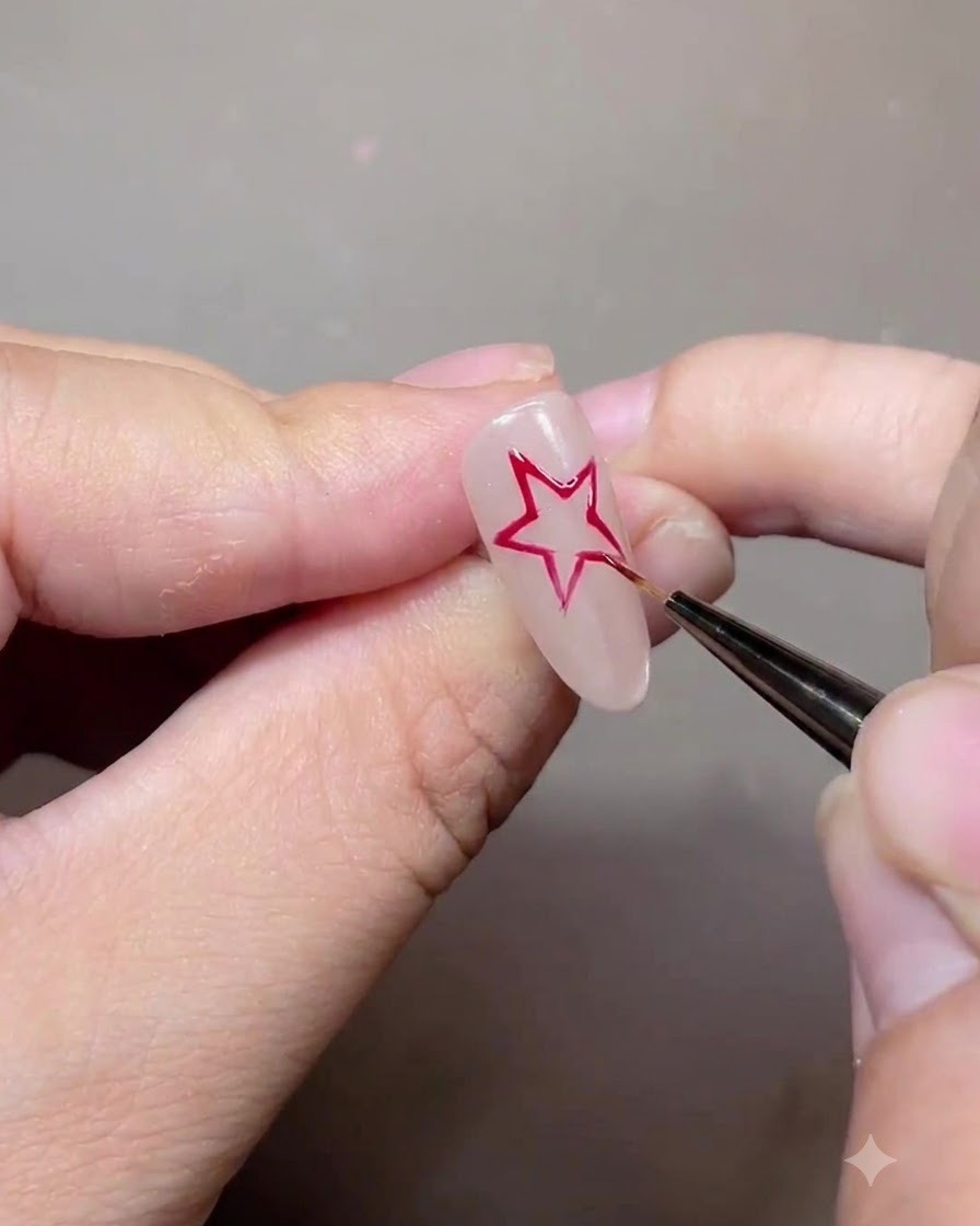 Ein feiner Liner-Pinsel zeichnet eine rote fünfzackige Sternumrisslinie auf einen Nude-Gel-Nagel.