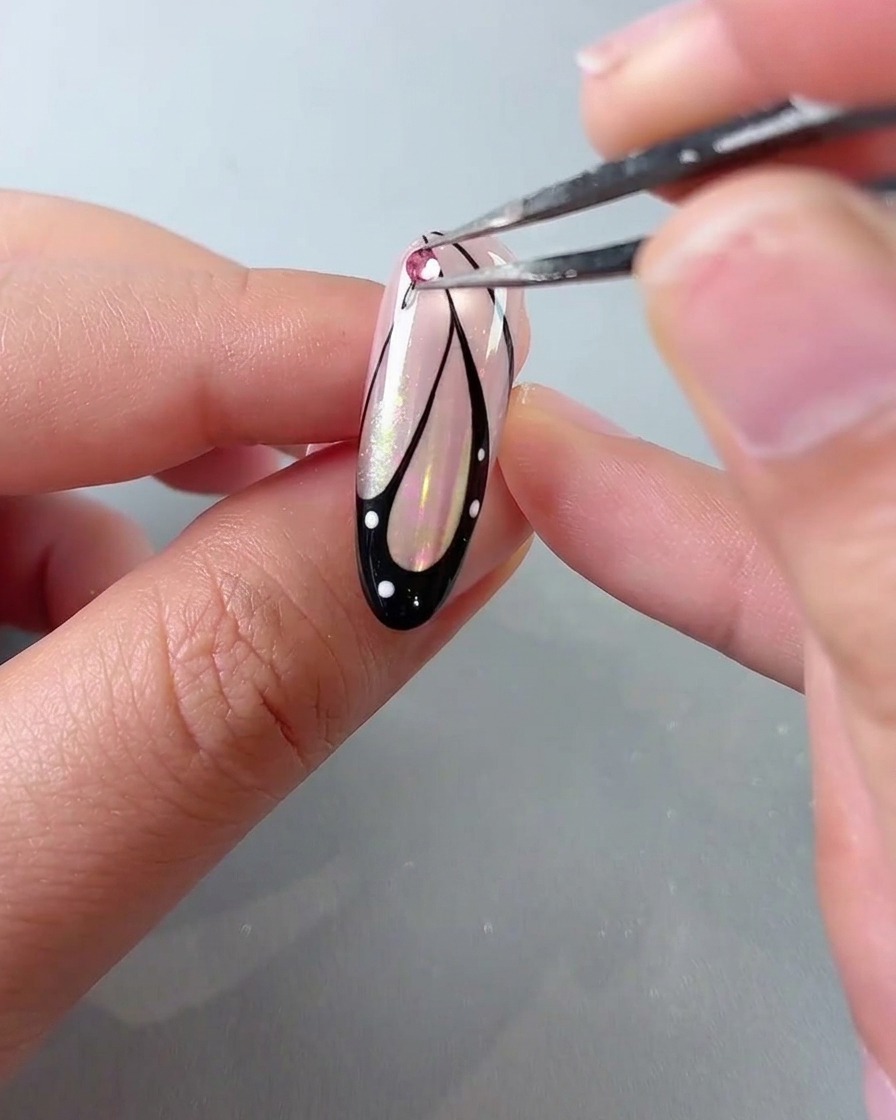 Eine Pinzette platziert einen rosa Kristall-Strassstein auf der Oberseite eines Schmetterlingsflügel-Nagelkunstdesigns.