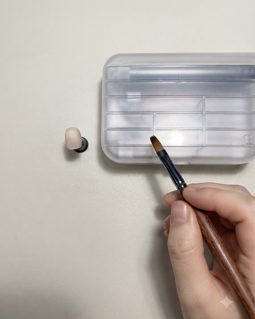 Eine Hand, die einen holzgefassten Nail-Art-Pinsel neben einem nudefarbenen Übungsnagel auf einem Ständer hält.