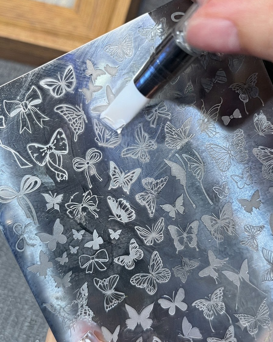 Ein Werkzeug verteilt weißes Stamping-Gel über ein Schmetterlingsmuster auf einer Metall-Stamping-Platte.