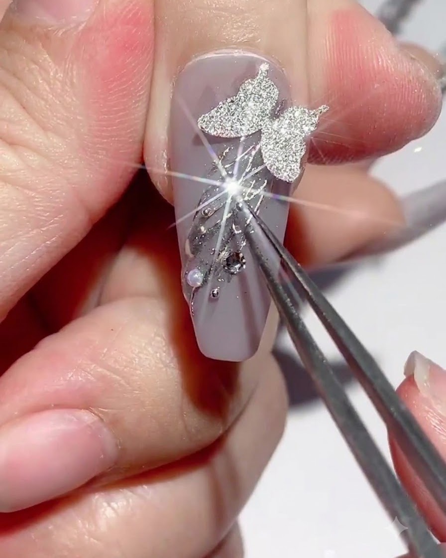 Eine Pinzette hält einen Charm, während Strasskleber um Nagelverzierungen herum aufgetragen wird.