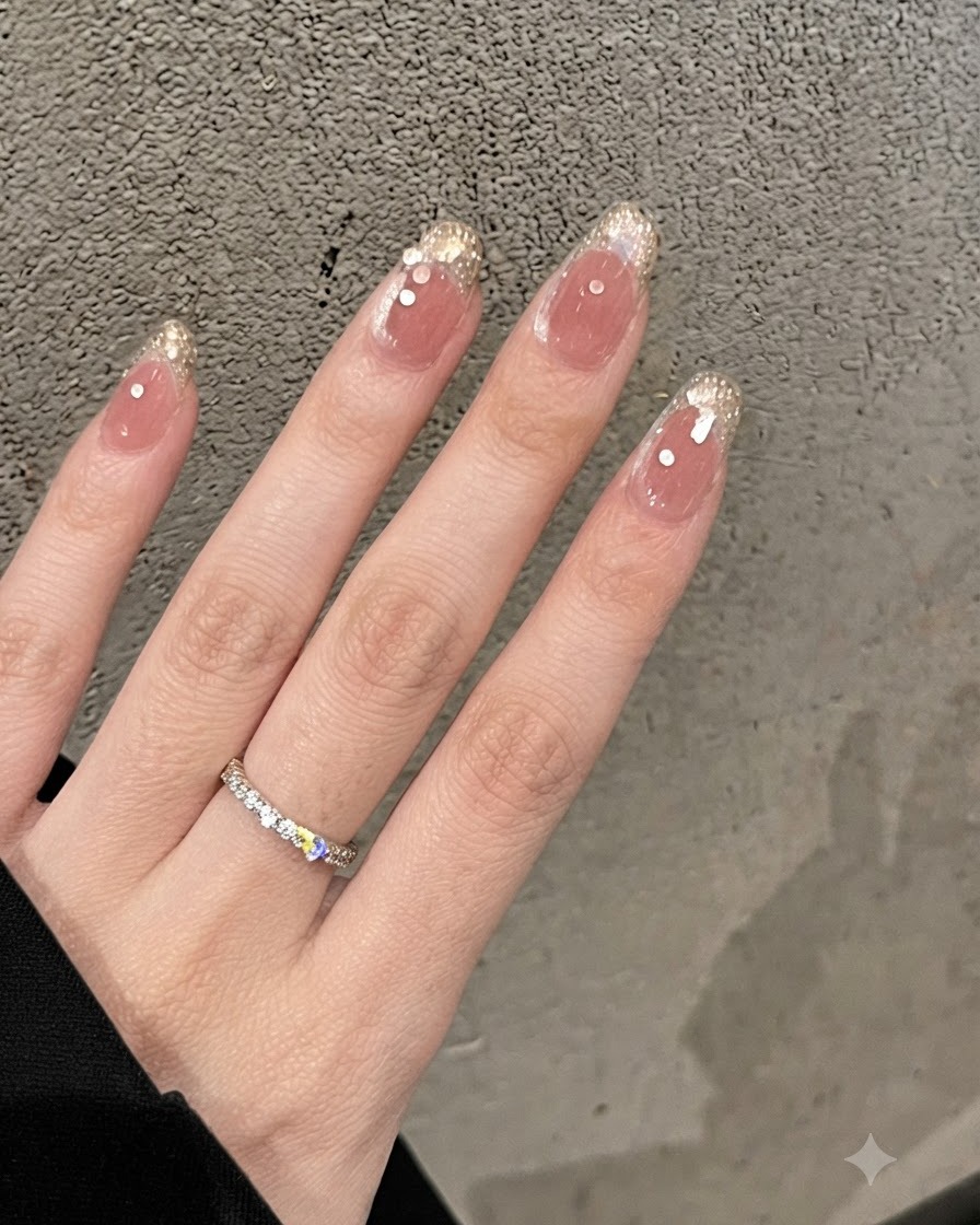 Nahaufnahme von Nude-farbenen Nägeln mit goldenen Glitter-French-Spitzen und winzigen Strasssteinen, mit einem Diamantbandring am Finger.