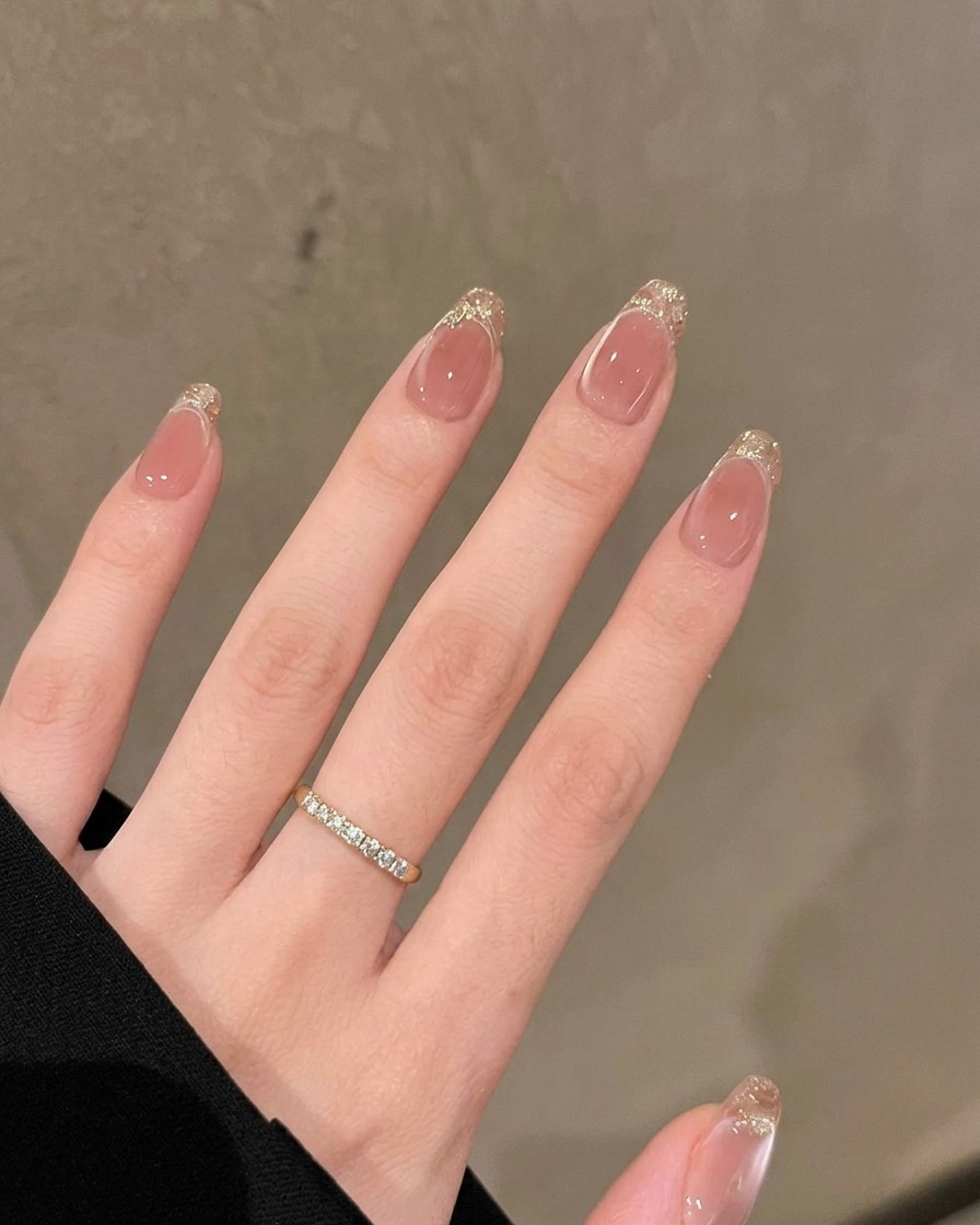 Schwenkaufnahme einer Hand, die lange Mandel-Nägel mit funkelnden goldenen Glitterspitzen und einem Hochglanz-Finish vor einer neutralen Wand zeigt.