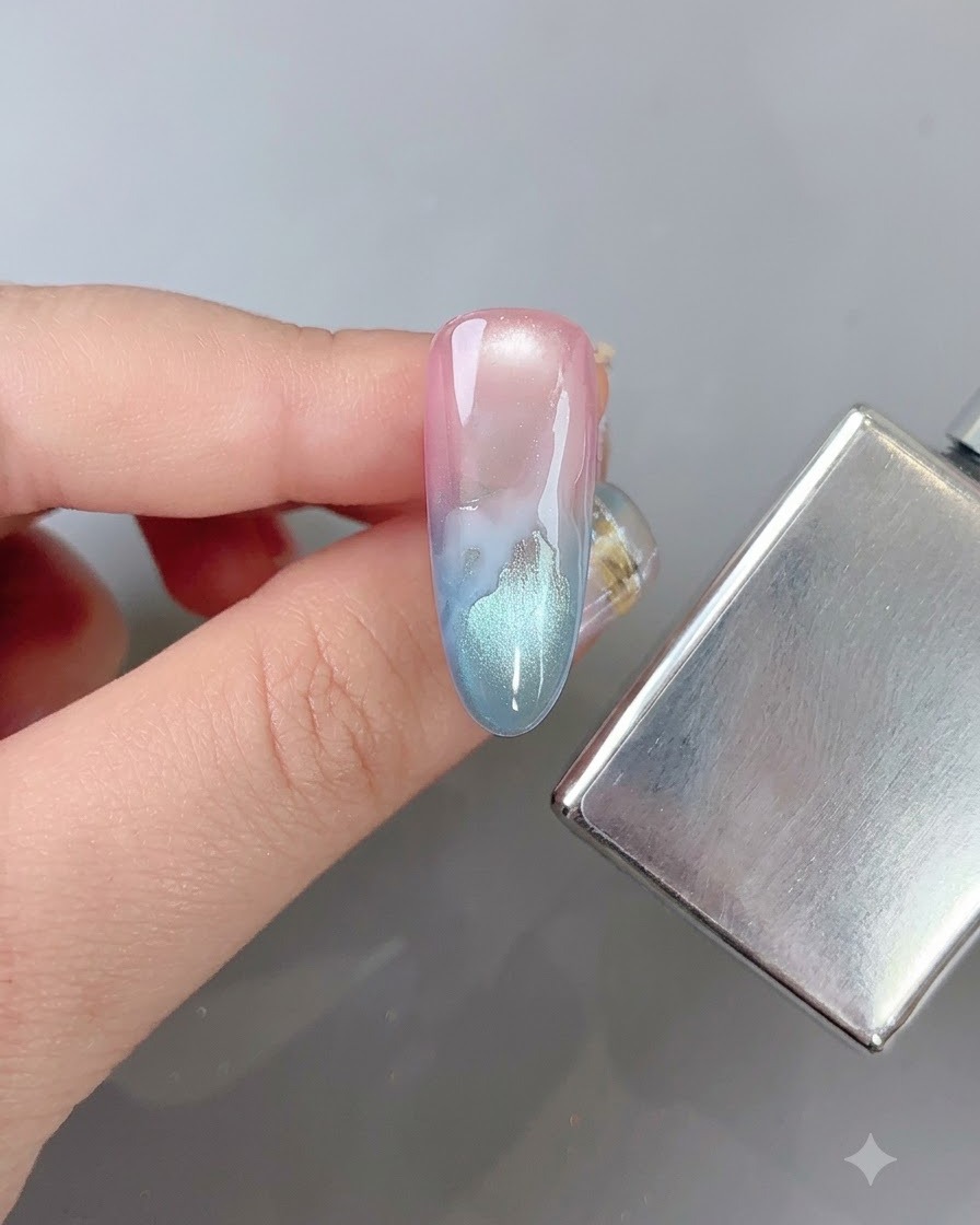 Ein Nagelpinsel verwischt weich die Ränder eines weißen Wolkenmusters auf einem mehrfarbigen Verlaufsnagel neben einem Magnetstick.