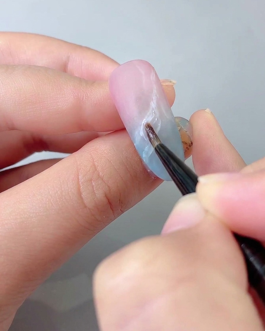 Ein feiner Detailpinsel zeichnet filigrane Schleifenlinien mit klebrigem Steinkleber auf die Nageloberfläche.