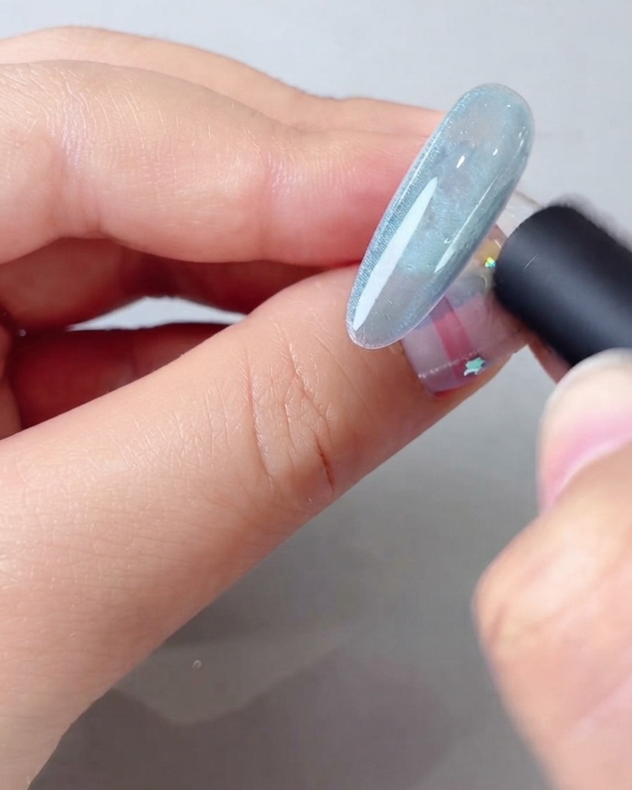 Ein Magnetwerkzeug wird nah an einen blauen Cat-Eye-Gel-Nagel gehalten, um einen breiten, schimmernden Lichtstrahl zu formen.
