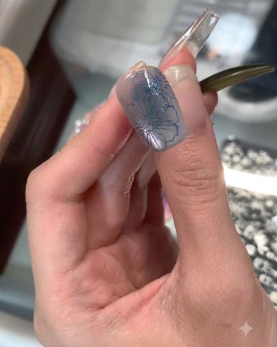 Ein klarer Silikonstempel trägt ein filigranes blaues Blumenstamping auf die Spitze eines Zeigefingernagels auf.