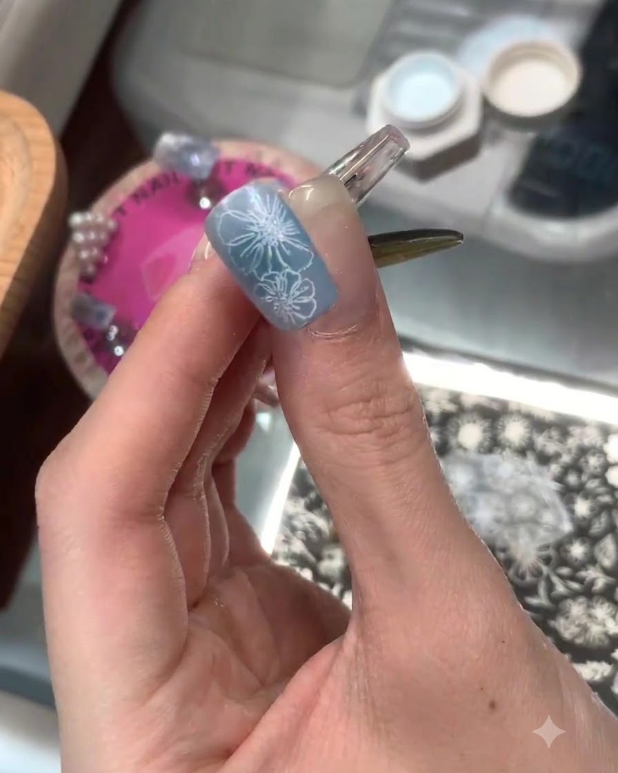 Ein manikürter Finger zeigt eine vollendete blaue Basis mit scharfen, weiß gestempelten Blumendetails auf der Nagelspitze.