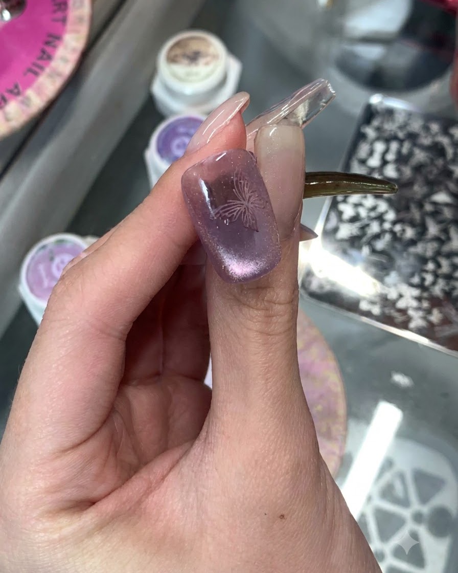 Eine Nahaufnahme eines lila Cat-Eye-manikierten Nagels mit einem filigranen gestempelten Schmetterlingsdesign darauf.