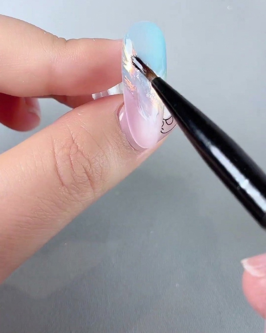Ein feiner Nail-Art-Pinsel trägt funkelnde, irisierende Glitzerdetails auf einen pastellfarbenen Ombré-Nagel auf.