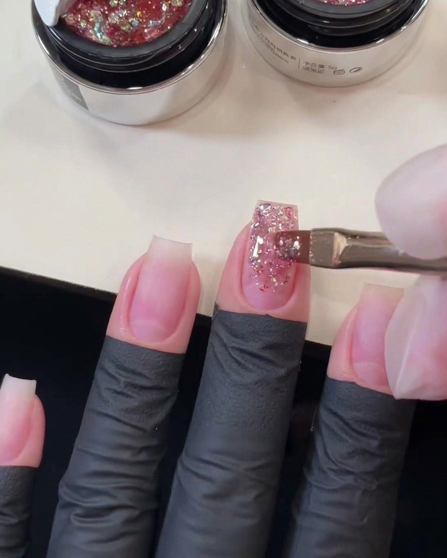 Flacher Gelpinsel, der rosa grobe Pailletten über die Länge des Mittelfingernagels zieht, um einen Glitzerverlauf zu formen.