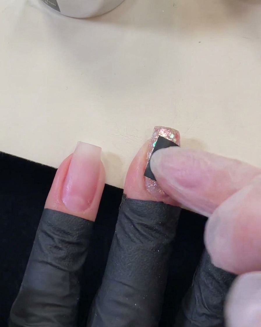 Ein Finger, der einen kleinen schwarzen quadratischen Aufkleber mit sternförmigem Ausschnitt auf einen glitzernden Paillettennagel drückt.