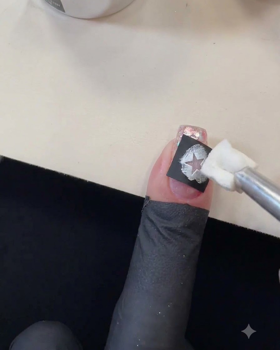 Ein Schwamm-Applikator, der weißes Gel-Polish in den Ausschnitt einer schwarzen Sternschablone auf einem glitzernden Nagel tupft.