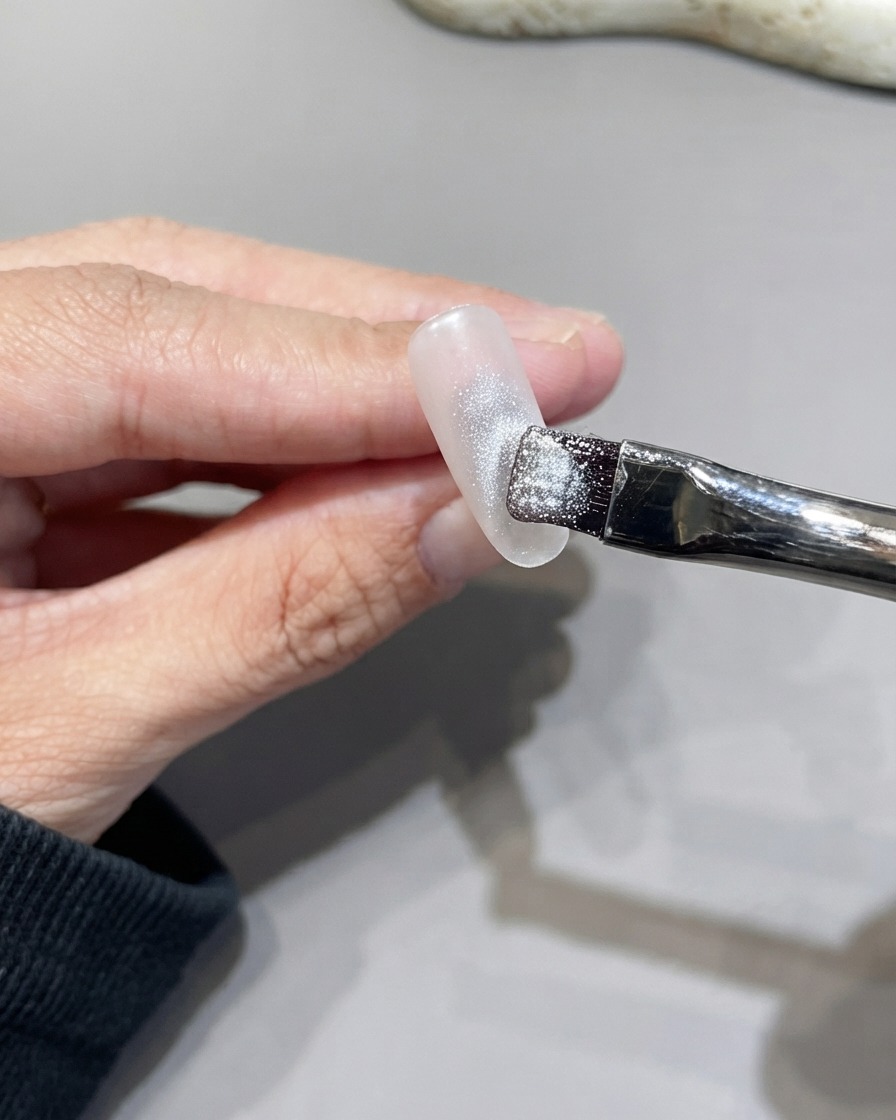 Ein flacher Pinsel trägt glitzerndes silbernes Cat-Eye-Gel gleichmäßig auf die Nageloberfläche auf.