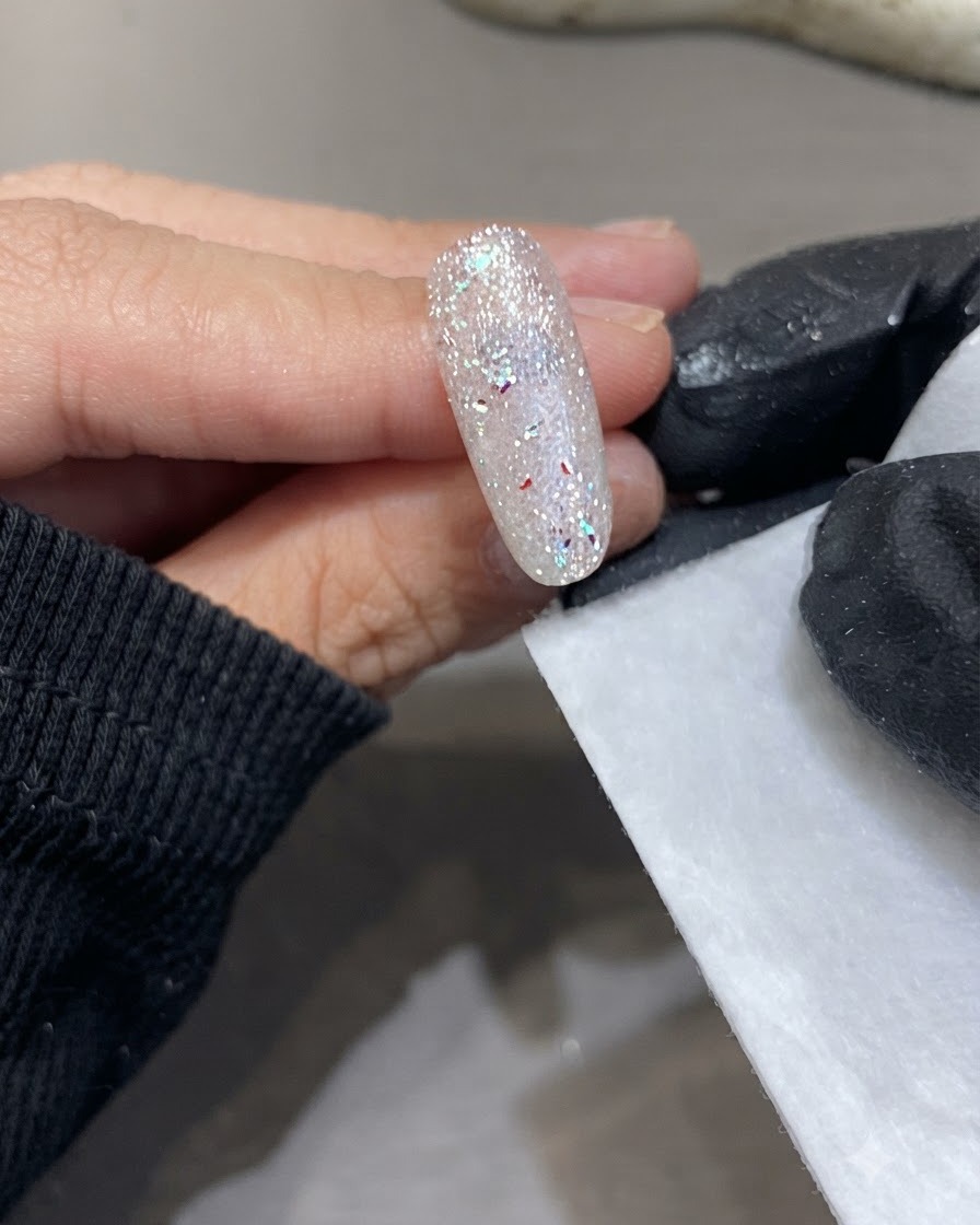 Eine bemalte Hand hält ein fusselfreies Tuch neben eine ausgehärtete, glitzernde Nageloberfläche.