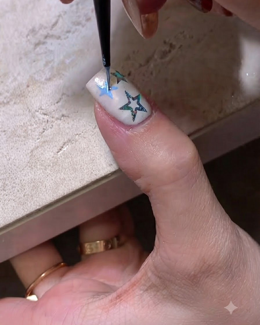 Nahaufnahme eines feinen Nail-Art-Pinsels, der einen blauen Sternumriss auf einen manikierten Nagel zeichnet.