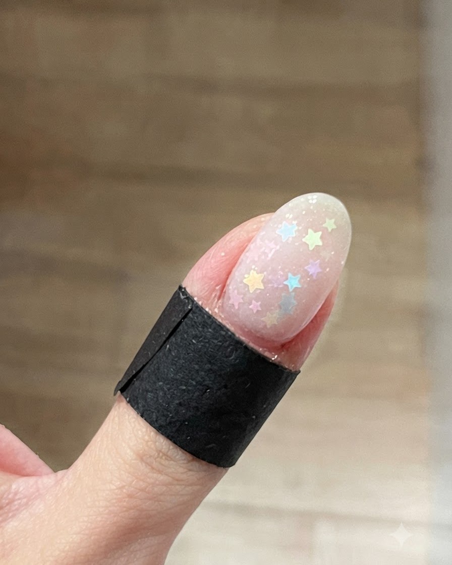Eine Nahaufnahme eines ovalen Fingernagels mit bunten Pastell-Stern-Glitzern, die auf einem klaren, feuchten Base Gel verstreut sind.