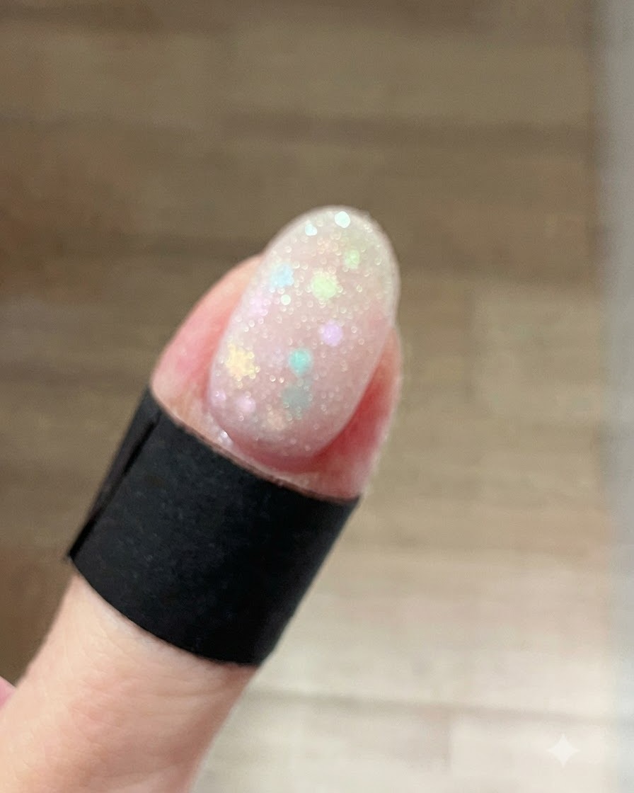 Eine Nahaufnahme eines Fingernagels, der mit einem nassen, funkelnden frostweißen Schimmergel über Pastell-Stern-Glitzern beschichtet ist.